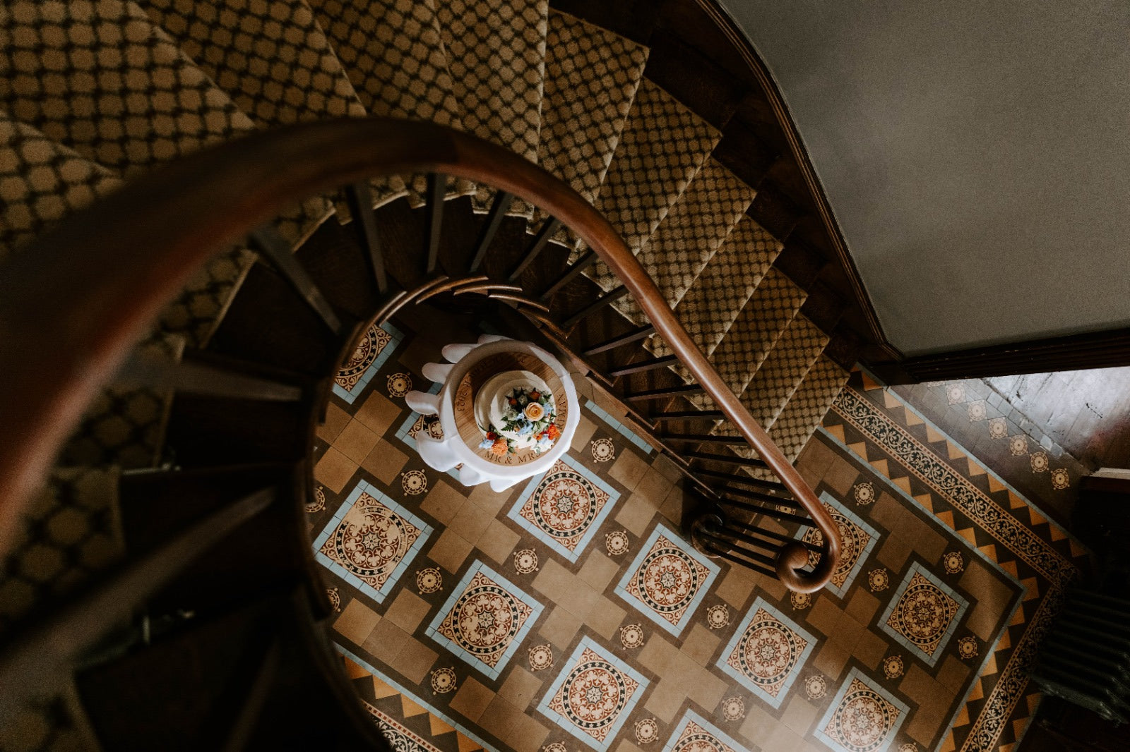 Spiral staircase with decorative patterned floor and a small table with a white cloth and floral arrangement with an oak cake board and white wedding cake.